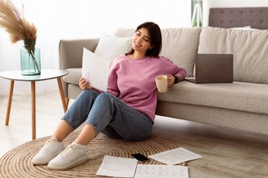 İçişleri Bakanlığı ve Teleworking Concept. Çalışan, elinde kağıt tutan ve mali belge okuyan gülümseyen genç bir kadın. Kadın işçi rapor veriyor, kahve içiyor, halının üzerinde oturuyor.