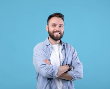 Kollarını kavuşturup mavi stüdyo arka planında kameraya gülümseyen pozitiflerin portresi. Sıradan giyinmiş, gülen yüzlü, maskülen sakallı, milenyum erkeği.