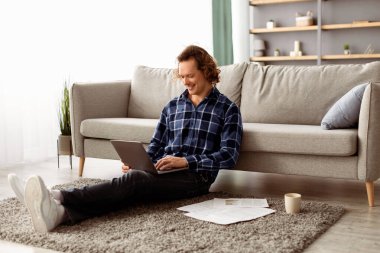 Uzak çalışma. Mutlu Serbest Çalışan Adam dizüstü bilgisayarını kullanıyor. Evde, yerde oturuyor. Oturma odasındaki bilgisayarda interneti kolaçan eden rahat adam. Bağımsız, Uzak İş, İnternet ve Teknoloji