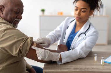 Close up of black female doctor in protective gloves applying sticking patch to patients arm after injecting elderly man with seasonal flu or infection vaccine, selective focus on hand clipart