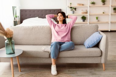 Dinlenme Konsepti. Oturma odasındaki rahat kanepede oturan güzel bayan, pencereye bakıyor. Mutlu kadın kanepede dinleniyor, arkana yaslanıyor, hafta sonunun keyfini çıkarıyor ya da işten çıkıyor.