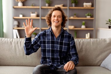 Merhaba. Merhaba. Evdeki koltukta oturmuş kameraya el sallayan neşeli adam. Gülümseyen ve el kol hareketi yapan genç adam içeride poz veriyor. Video Çağrısı, Modern Uzaklık İletişim Kavramı