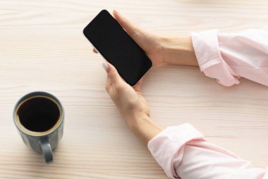 Yüksek açılı bir bakış açısıyla, tasarım için siyah ekranı olan akıllı telefonu kullanan, elinde tutan ve kullanan bir kadın, Breskfast içen, sabah kahve içen, internette yemek tarifleri arayan, bedava alan arayan,