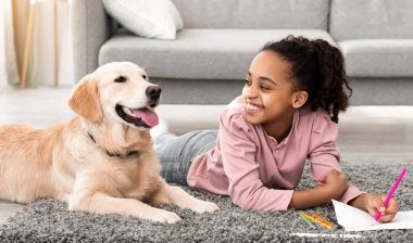 Çocuklar ve Evcil Hayvan. Neşeli zenci kız portresi köpeğiyle yerde yatar renkli pastel boyalarla resim çizer oturma odasında, pankartta, pankartta altın emekliye bakar.