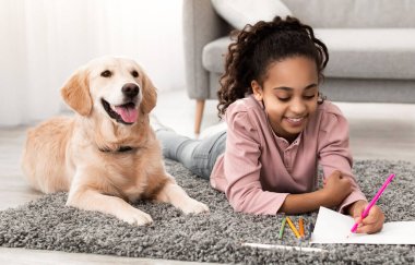 Çocuk ve Aile Hayvanları Konsepti. Oturma odasında labradoruyla halıya uzanmış, renkli boya kalemleri ve kalemlerle resim çizen gülümseyen zenci kızın portresi.