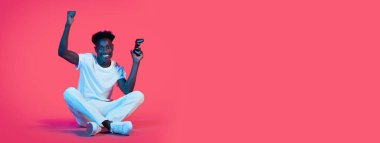 Genç siyah bir adam yerde bağdaş kurmuş oturuyor, video oyunu kontrolörlerini tutarken heyecanla kollarını kaldırıyor..