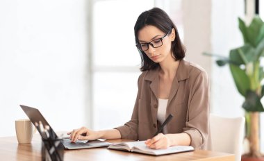 Not alıyorum. Dizüstü bilgisayar kullanan ve modern ofisteki günlüğüne not yazan bir kız öğrencinin portresi.