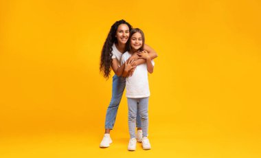 Orta Doğulu Anne, sarı arka planda stüdyoda duran küçük kızına sarılıyor. Arap anne ve kızı kameraya sarılıp gülümsüyor. Anneliğin neşesi