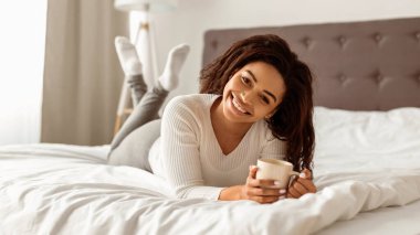 Günün iyi başlangıcının tadını çıkarıyorum. Beyaz çarşaflarla rahat bir yatakta dinlenen kıvırcık saçlı güzel Afro-Amerikan kadın portresi. Pozitif kadın poz veriyor ve kameraya bakıyor, kahve içiyor.