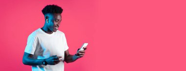 Genç bir siyah adam renkli bir stüdyoda duruyor, akıllı telefonuyla müzik dinlerken gülümsüyor. Kablosuz kulaklık takıyor ve elinde bir cihaz tutuyor..
