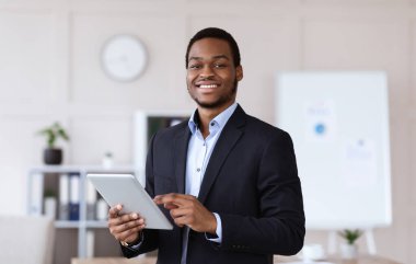 Gülümseyen siyah girişimci genç adam dijital tablet tutuyor, modern teknolojileri iş dünyasında, ofis içinde, kopyalama alanında kullanıyor. Neşeli Afro-Amerikan iş adamı iş için uygulama kullanıyor.