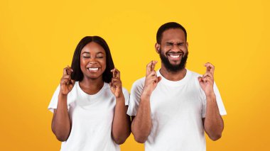 Değer Veren Dilek. Pozitif siyah çift parmaklarını çapraz yapıp gülümsüyor, şans diliyor, mucize için dua ediyor, iyi şeyler olmasını umuyor, sarı stüdyonun duvarında kapalı gözlerle dikiliyor.