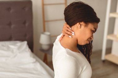 Closeup of irritated African American young woman suffering from pain in body after sleeping, touching and massaging her neck, having uncomfortable bed and mattress, home interior, free copy space clipart