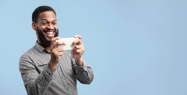 Gadget Bağımlılık Konsepti. Cep telefonuyla oyun oynayan mutlu siyahi adamın portresi mavi stüdyo arka planında izole edilmiş, kopyalama alanı. Onun cihazını kullanarak video izleyerek tatmin olmuş bir erkek.