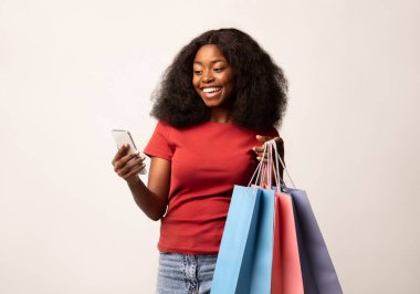 Çevrimiçi alışveriş için akıllı telefondaki mobil uygulamayı kullanan mutlu siyahi kadın, neşeli Afrikalı Amerikalı kadın, parlak çantalar ve cep telefonları tutuyor, sezonluk satışların ve mobil tekliflerin tadını çıkarıyor, kopyala