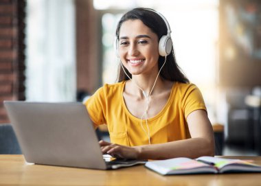 Yüzünde kulaklıkla gülümseyen Arap kız internette ders çalışıyor, kafede dizüstü bilgisayar kullanıyor, notlar alıyor, yer kopyalıyor.