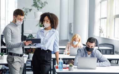 Meslektaşların iş yerindeki hatalar hakkında tartışması. Guy ve koruyucu maskeli Afrikalı Amerikalı kadın, kitle imha silahı salgını sırasında işçilerle birlikte modern ofis içinde tablete bakıyor ve konuşuyorlar.