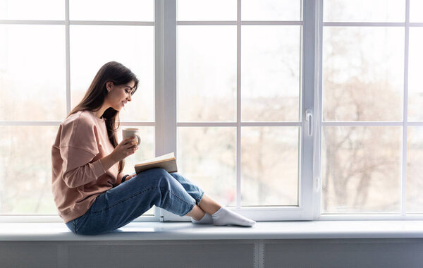 Free Time To Relax. Profile of beautiful woman sitting on white windowsill at home, reading paper book, holding cup and drinking hot coffee. Happy lady taking break with mug near window, copy space