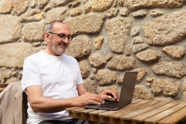 Olgun bir adam, sevimli taş bir duvarın olduğu samimi bir kahve dükkanında laptopuna odaklanmış, kırsal ahşap bir masada oturuyor. Çalışırken meşgul ve memnun görünüyor..