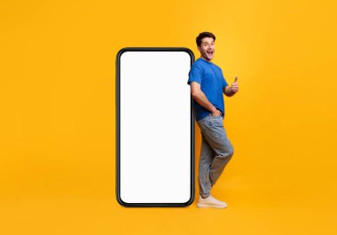 Neşeli genç adam beyaz ekranlı, kocaman, boş bir cep telefonuna yaslanmış ve el hareketini gösteriyor. Model, sıradan adam havalı mobil uygulama öneriyor. Parlak sarı arka plan, tam boy.