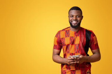 Gülümseyen siyah adam modern akıllı telefon kullanıyor, Afrika tişörtü giyiyor, sarı arka plan