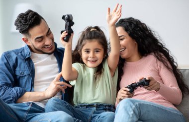 Evde video oyunu oynayan üç kişilik mutlu ortadoğu ailesi, genç Arap ebeveynler ve küçük kızları oturma odasında eğleniyorlar, ellerinde joysticklerle gülüyorlar, yakın çekim yapıyorlar.