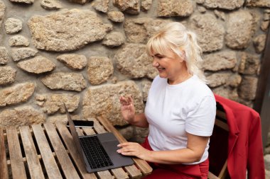 Tahta bir masada oturan bir kadın, temiz havanın tadını çıkarırken dizüstü bilgisayarına odaklanmış. Arkasındaki taştan duvar cazibesini arttırıyor. Öğleden sonraki görevleri için sakin bir çalışma alanı yaratıyor..