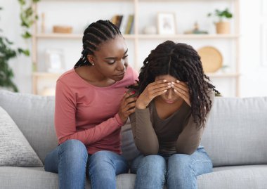 Attentive african american lady comforting her upset crying girlfriend or sister, giving her hug, saying supportive words, living room interior, copy space. Support in friendship concept