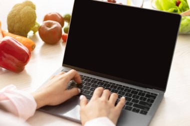 Omuz hizasında genç bir kadın siyah boş ekranlı dizüstü bilgisayar kullanıyor, mutfakta yemek yapıyor, klavyede yazıyor, internet üzerinden vegan tarifleri arıyor.