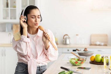 Mutfak eğlencesi. Müzik dinleyen, en sevdiği şarkıyı söyleyen, mikrofon olarak tahta spatula kullanan kablosuz kulaklık takan ve müzik eşliğinde dans eden genç bir bayan.
