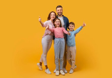 Anne, baba, oğul ve kızdan oluşan mutlu bir aile birbirine yakın duruyor, hepsi de işaret veriyor. Canlı sarı bir arkaplanda poz veriyorlar.