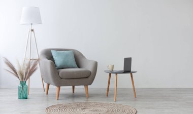 Uzaktan çalışma ve evde rahatlık için rahat bir boho tasarımı. Yastıklı gri eski koltuk, kahve ve dizüstü bilgisayarlı masa, kuru bitkilerle dolu vazo, yerde lamba ve halı, arka planda ışık duvarı