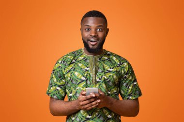 Afrika kıyafetleri içinde heyecanlı bir zenci modern cep telefonu tutuyor, yeni uygulama indiriyor, sarı arka plan