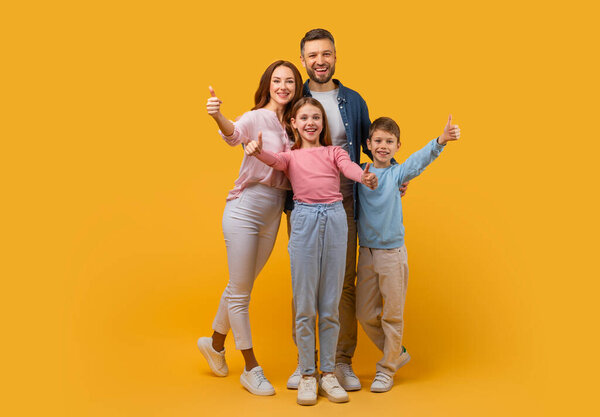 Happy family consisting of a mother, father, son, and daughter stands close together, all giving a thumbs up sign. They are posing against a vivid yellow backdrop