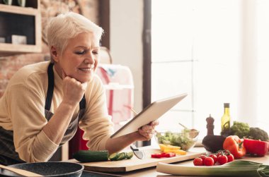 İnternetteki Tarifler. Mutfakta dijital tablet kullanan yaşlı bir kadın internetteki malzemeleri kontrol ediyor.