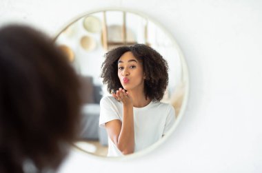 Harika görünüş, kendini ve mükemmel cildini sev. Neşeli Afro-Amerikan bayan hava öpücüğü veriyor ve aynanın yansımasına bakıyor. Oturma odasında, beyaz duvarda, panoramada, boş alanda.