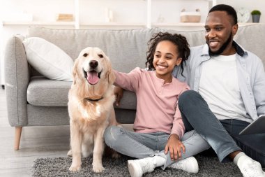 Mutlu Afrika kökenli Amerikalı baba ve kız oturma odasındaki halıda oturuyor, evcil köpekleri ile vakit geçiriyorlar, olumlu adam tablet tutuyor, neşeli kız evcil hayvanlarını okşuyor.