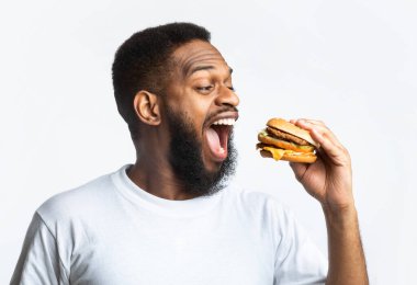 Beyaz arka planda stüdyoda durup hamburger yiyen Afro-Amerikalı adamın yan görüntüsü. Abur cubur ve aşırı yeme alışkanlığı. Zenci Isıran Çizburger Sağlıksız Beslenme Konsepti