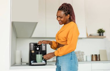 Excited young black woman making fresh aromatic coffee in modern machine in kitchen interior, enjoying hot morning beverage at home, free space