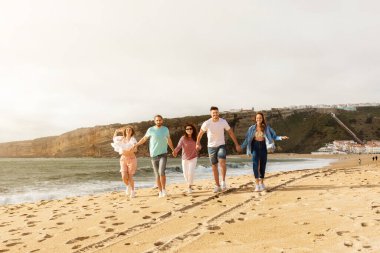 Beş arkadaştan oluşan bir grup kumsalda el ele yürüyor, gülüyor ve güneşli bir günün tadını çıkarıyorlar. Okyanus dalgaları arka planda yavaşça kıyıya vurur..