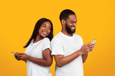 Halk ve Teknoloji Konsepti. Sırt sırta verip cep telefonlarını tutan ve kullanan neşeli Afro-Amerikan çiftin portresi, erkek arkadaşına bakan gülümseyen zenci kadın.