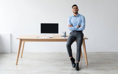 Şablonu Uygula. Masada oturan mutlu, gülümseyen bir Arap, kolları kavuşturulmuş bir şekilde poz veriyor. Bilgisayarı merkez büroda boş bir siyah ekranla gösteriyor. Bedava kopya alanı. İnsanlar, Teknoloji, Uzaktan Çalışma Konsepti