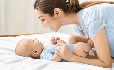 Bebekler için konuşma ve dil gelişimi. Genç anne, yeni doğmuş bebeğiyle yatakta, yan gözle oynuyor.