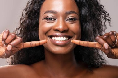 Diş hizalama ve beyazlatma konsepti. Mutlu Afrikalı Amerikalı kız, gri arka planda iki işaret parmağıyla geniş gülüşünü işaret ederek, yakın plan