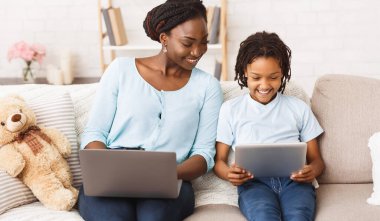 Evde kal Afrikalı anne uzaktan bilgisayarla çalışıyor ve çocuğuna ödevlerinde yardım ediyor, tablet ve laptop kullanıyor.
