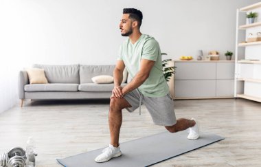 Formda genç bir Arap hamlesini yapıyor, güç egzersizleri yapıyor, evde bacak kaslarını çalıştırıyor, tam boy. Milenyum Doğulu adam idman yapıyor, ev içi spor yapıyor.