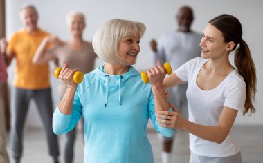 Kol halterleriyle egzersiz yaparken aktif kıdemli kadınlara yardımcı olan kadın fitness koçu, huzurevinde beden eğitimi alan ve eğitmenle eğitim gören çok ırklı spor grubu.