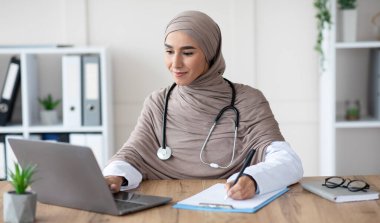 İnternette randevusu olan genç bir bayan doktor, ofiste oturuyor, dizüstü bilgisayar kullanıyor ve notlar alıyor, fotokopi çekiyor. Çekici Müslüman kadın doktor bilgisayar, klinik içi, uzaktan sağlık hizmetleri ile çalışıyor.