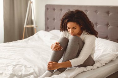 Depresyon Konsepti. Erkek arkadaşı ya da kocasından ayrıldıktan sonra yatakta yalnız oturan ve bacakları kucaklaşan mutsuz bir Afro-Amerikan kadının portresi. Üzgün kadın kendini yalnız hissediyor