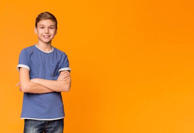 Sevimli genç çocuk kendinden emin hissediyorum, katlanmış ellerle poz ve turuncu stüdyo arka plan, panorama üzerinde gülümseyerek